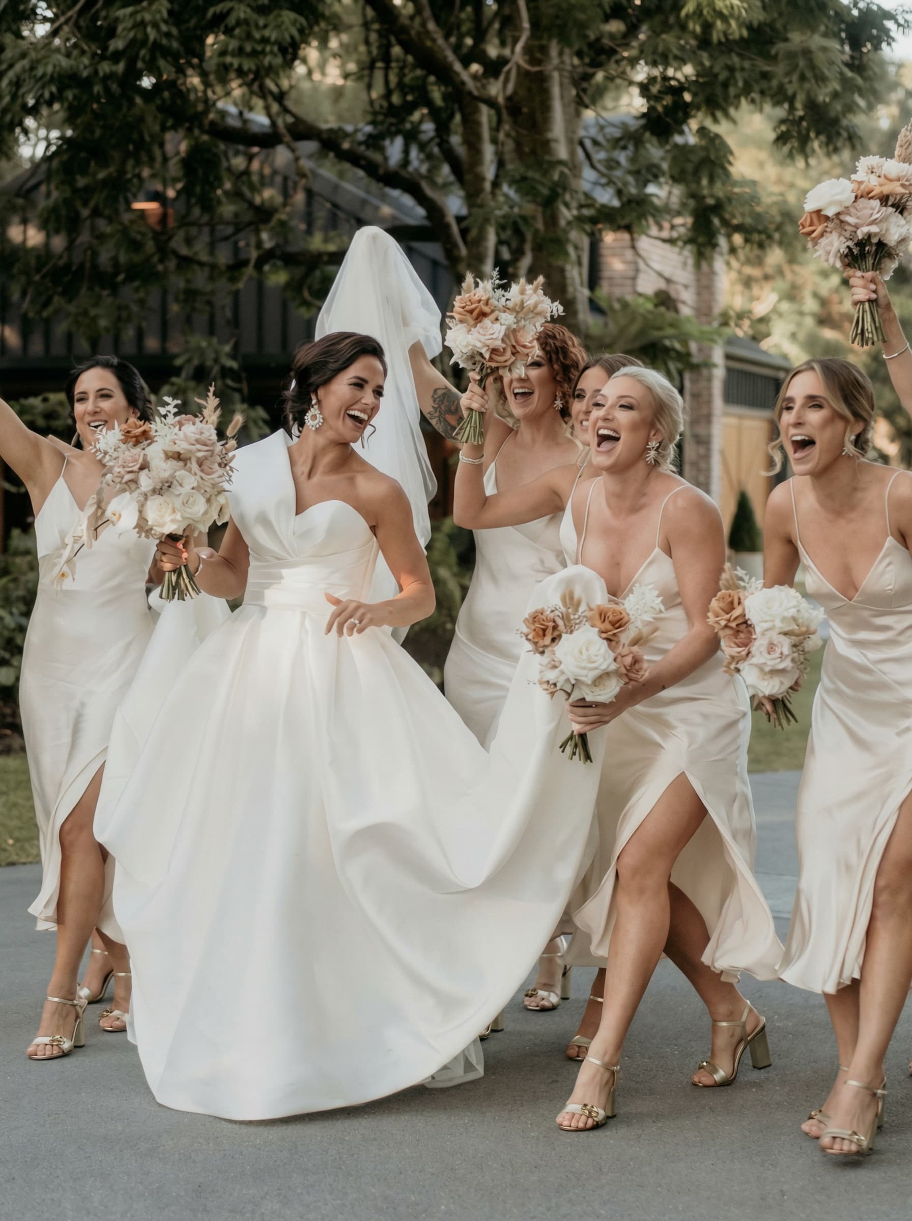 A group of girls following a bride and holding flowers and expressing happness and wearing white maxi dresses 