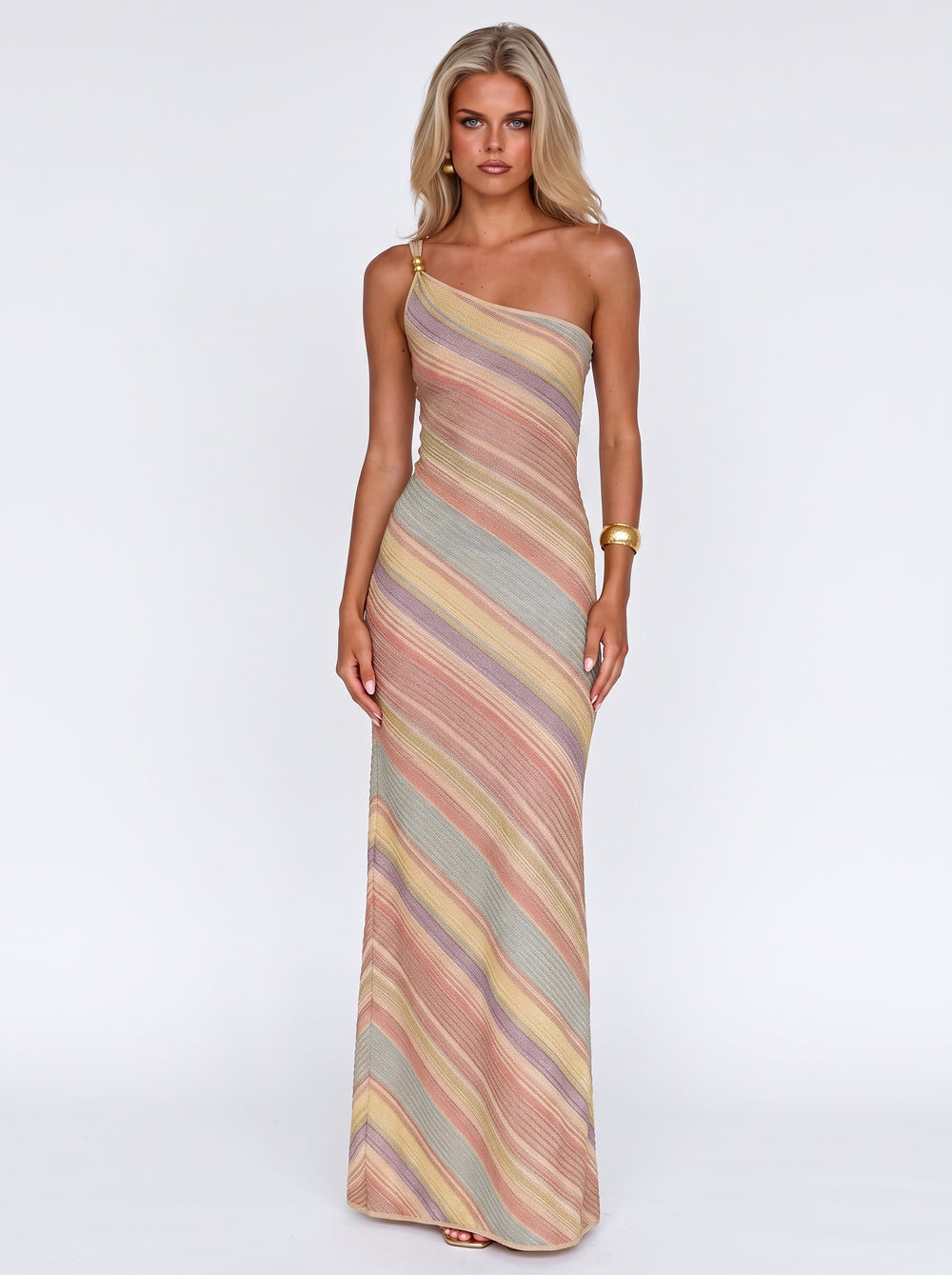 A woman modeling a one-shoulder maxi dress with diagonal stripes in muted pink, beige, yellow, and gray tones against a plain white studio background.