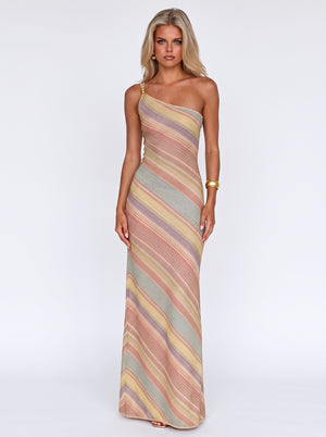 A woman modeling a one-shoulder maxi dress with diagonal stripes in muted pink, beige, yellow, and gray tones against a plain white studio background.