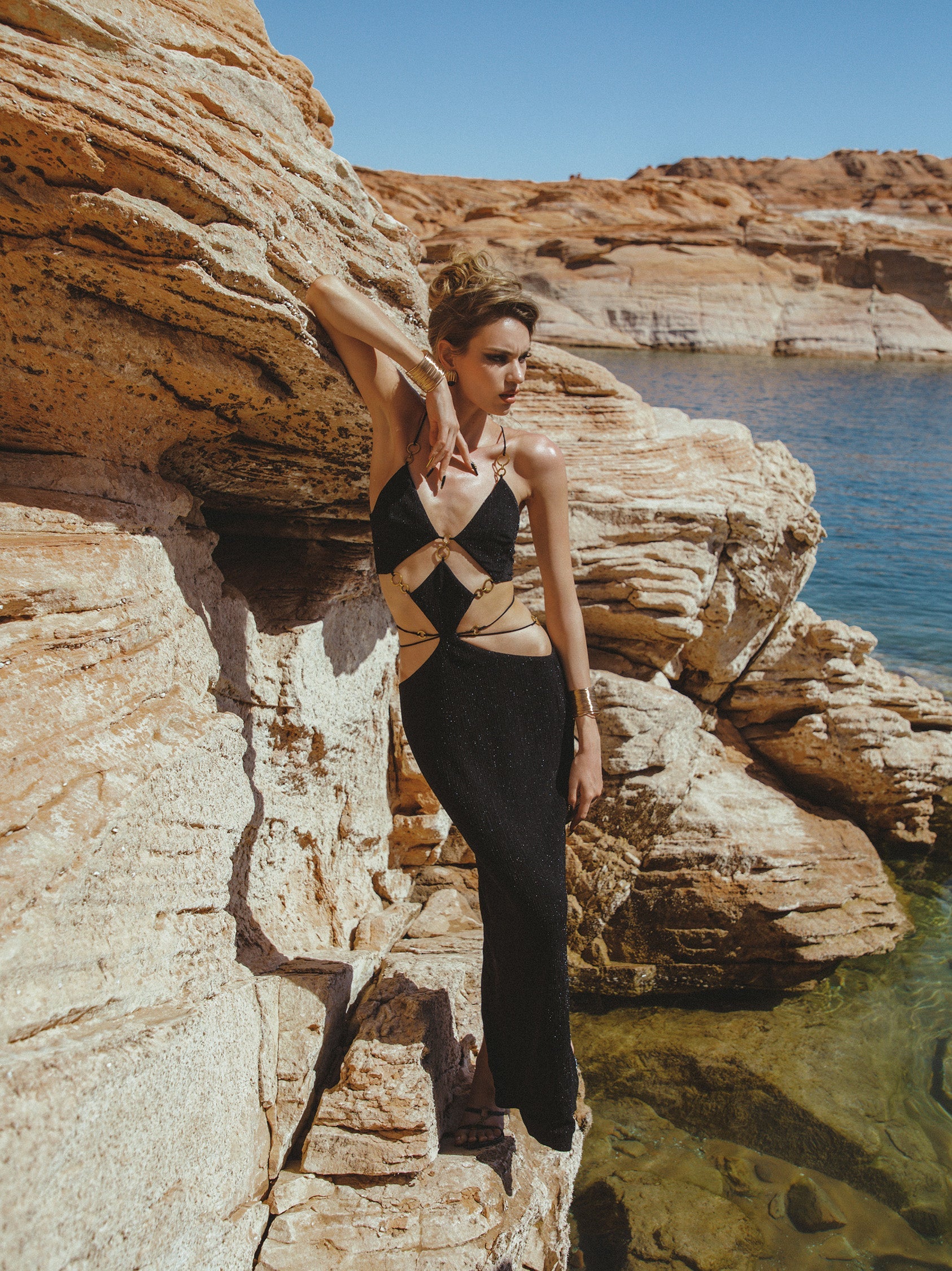 Woman in a black and white patterned top and black pants standing on rocky cliffs with water and sky in the background.