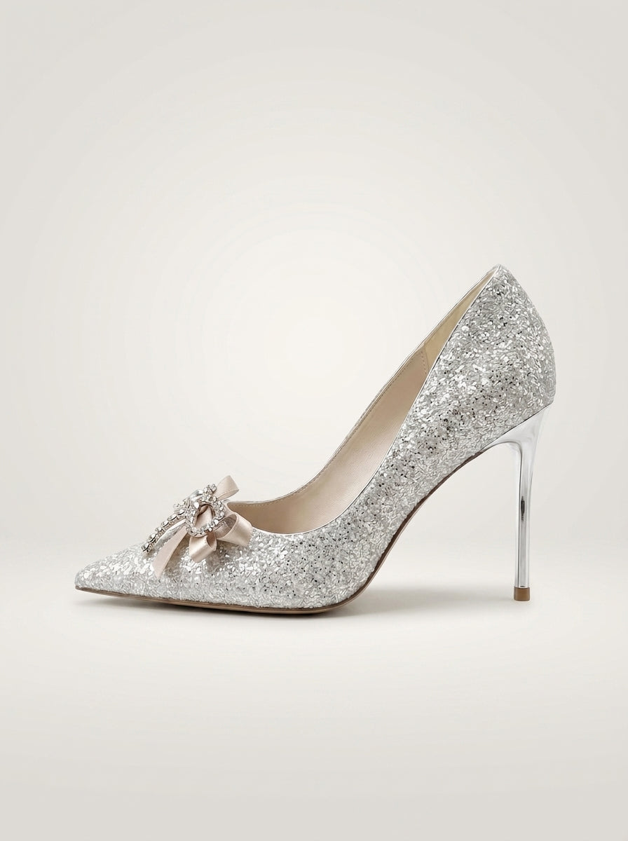 A single silver glitter pointed-toe pump with a slim high heel and a small pale pink bow embellishment on the toe, shown against a plain light beige studio background.