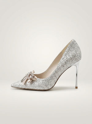 A single silver glitter pointed-toe pump with a slim high heel and a small pale pink bow embellishment on the toe, shown against a plain light beige studio background.