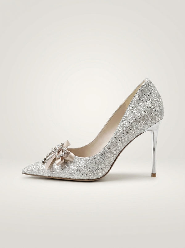 A single silver glitter pointed-toe pump with a slim high heel and a small pale pink bow embellishment on the toe, shown against a plain light beige studio background.