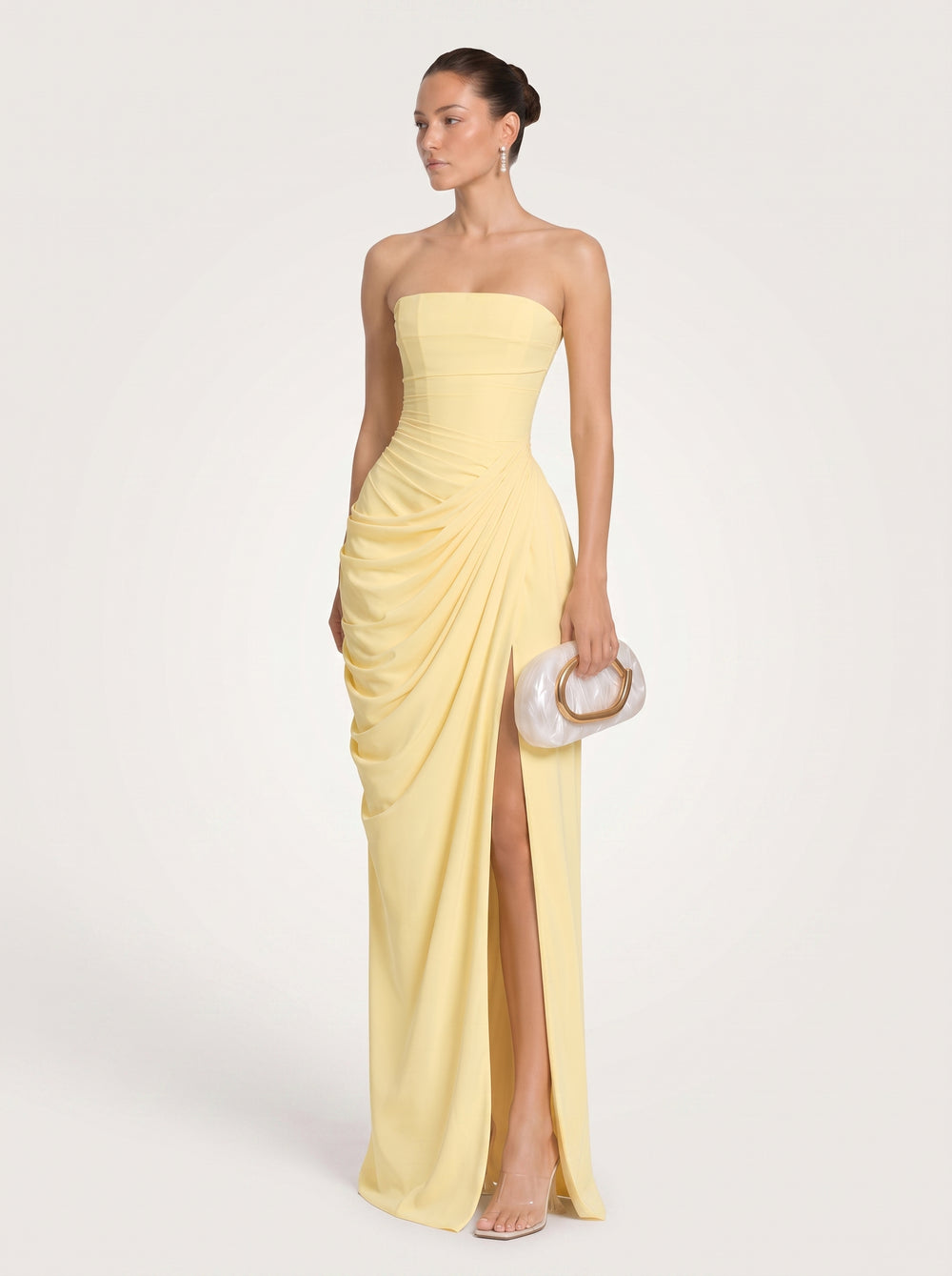 A woman modeling a strapless pale yellow ruched dress with a draped skirt and thigh-high slit, holding a white clutch with a gold ring handle against a light beige studio background.